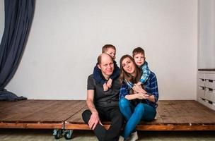 familia sonriente sosteniendo la forma de la casa mientras se sienta en el piso de madera dura en casa. sesión de fotos de una familia con 2 hijos, fondo blanco. cálidas relaciones familiares. padres jovenes