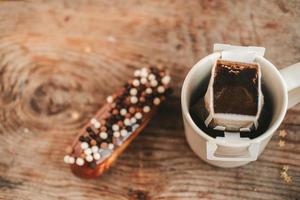 café en una bolsa de goteo en una taza blanca y un eclair con bolas sobre un fondo de madera, vista superior foto