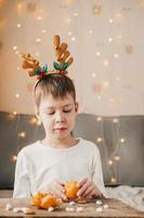 boy in Christmas antlers, sits and peels a tangerine on the background of a yellow garland. Boy in a white t-shirt at home, peels a tangerine on a wooden table with marshmallows photo