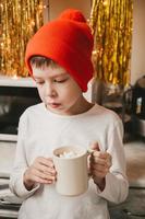 boy in a white t-shirt and a red hat drinking cocoa with marshmallows in the kitchen. Christmas mood. Christmas sweets and drinks photo