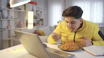 Obese boy eats pasta while watching video. The child with a weight problem eats pasta between classes and watches videos on the laptop.