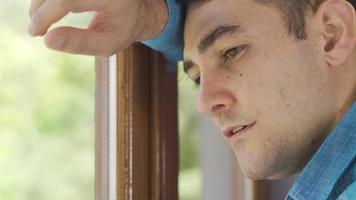 el hombre que apoya la cabeza contra la ventana mira tristemente por la ventana. hombre pensativo y aburrido mirando por la ventana. video