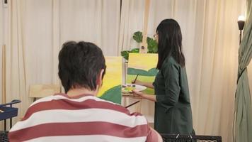 Female Asian teacher teaches and demonstrates to the student boy on acrylic color picture painting on canvas in art classroom, creatively learning with skill at the elementary school studio education. video