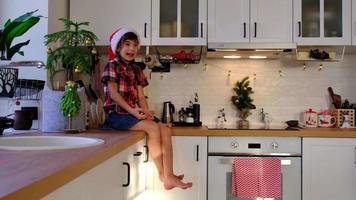 enfant dans un bonnet de noel est assis dans une cuisine festive décorée pour noël. fille heureuse grimaces drôles et joue avec des balles. nouvel An video