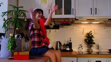 niña feliz está indignada, jura y amenaza con el puño que la caja está vacía y allí y muecas graciosas. niño con sombrero de santa está sentado en una cocina festiva decorada para navidad. Año Nuevo video