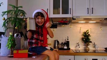 Happy girl is indignant, swears and threatens with his fist that box is empty and there  and funny grimaces. Child in a Santa hat is sitting in a festive kitchen decorated for Christmas.  New Year video