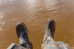 piernas de un pescador sentado en botas de goma sucias en la orilla del río con agua sucia en el fondo foto