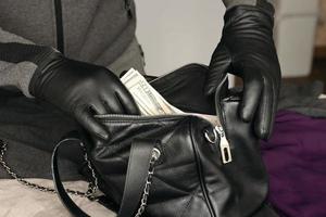 ladrón con traje negro y guantes que se ven en una bolsa de mujer robada abierta. el ladrón saca billetes de dólares estadounidenses del bolso de una mujer en la cocina foto