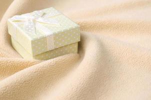 A small gift box in orange with a small bow lies on a blanket of soft and furry light orange fleece fabric with a lot of relief folds. Packing for a gift to your lovely girlfriend photo