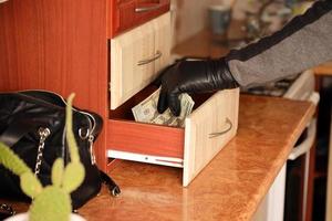 ladrón con traje negro y guantes se ve en un estante abierto en la cocina. el ladrón saca los billetes de dólar estadounidense de un estante foto