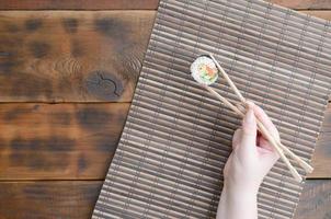 A hand with chopsticks holds a sushi roll on a bamboo straw serwing mat background. Traditional Asian food photo