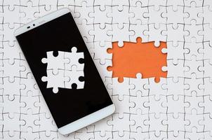 A modern big smartphone with several puzzle elements on the touch screen lies on a white jigsaw puzzle in an assembled state with missing elements photo