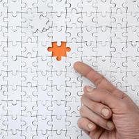 The texture of a white jigsaw puzzle in the assembled state with one missing element, forming an orange space, pointed to by the finger of the male hand photo