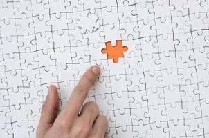 The texture of a white jigsaw puzzle in the assembled state with one missing element, forming an orange space, pointed to by the finger of the male hand photo