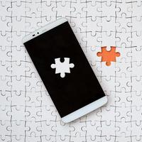 A modern big smartphone with several puzzle elements on the touch screen lies on a white jigsaw puzzle in an assembled state with missing elements photo
