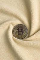 The golden bitcoin lies on a blanket made of soft and fluffy light orange fleece fabric with a large number of relief folds. The shape of the folds resembles a fan from a video card cooler photo