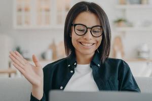 Smiling woman in glasses waving hand hello, answers video call, chatting online on laptop at home photo