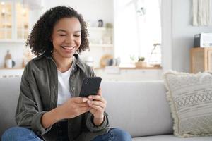 Happy mixed race girl uses mobile phone apps watching funny video in social networks sitting on sofa photo