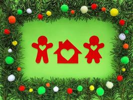 Gingerbread man family and their dream house cut out of red felt in frame made of fir tree branches around on light green background. Border decorated colorful pom poms. Christmas and New Year concept photo