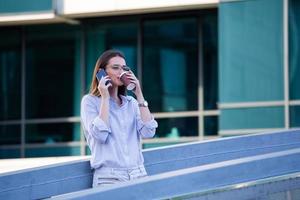 Executive business woman talking on mobile smartphone and drinking coffee from disposable paper cup in the street with office buildings in the background photo