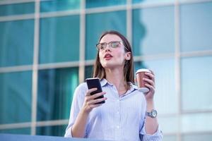 Executive business woman looking at mobile smartphone and drinking coffee from disposable paper cup in the street with office buildings in the background photo