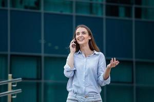 Happy smiling business woman talking on mobile smartphone and walking in the street with office buildings in the background photo