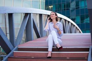 mujer de negocios sonriente feliz hablando en un teléfono inteligente móvil en la calle con edificios de oficinas en el fondo foto