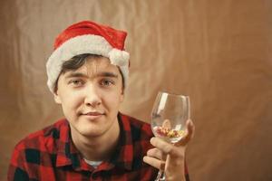 Guy in Santa Claus hat holds a glass with various pills. photo