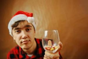 Guy in Santa Claus hat holds a glass with various pills. photo