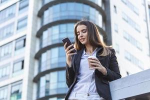 confident businesswoman texting on her mobile phone while drinking coffe in front of glass window of skyscraper building dreaming of success thinking of future business vision and new goals photo