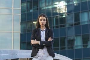 Successful female office worker with net-book is standing in skyscraper interior against big window with city view on background. Proud woman architect looking satisfied with completed project photo