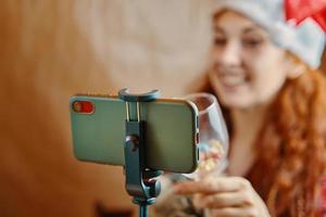 Red-haired girl in Santa Claus hat with glass in front of mobile phone. photo