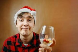 Guy in Santa Claus hat holds a glass with various pills. photo