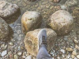 A foot in a sneaker steps on large round beautiful natural stones cobblestones in water, sea, lake, river. Concept tourism, outdoor activities photo