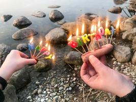 inscripción de feliz cumpleaños en llamas hecha de velas navideñas en manos de un hombre y una mujer frente al agua del río del lago oceánico. concepto de celebración de cumpleaños en la naturaleza, al aire libre foto