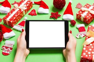 Top view of woman holding tablet in her hands on green background made of Christmas decorations. New Year holiday concept. Mockup photo