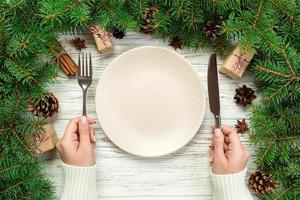 top view girl holds fork and knife in hand and is ready to eat. Empty plate round ceramic on wooden christmas background. holiday dinner dish concept with new year decor photo