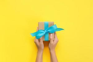 Woman hands give wrapped valentine or other holiday handmade present in paper with blue ribbon. Present box, decoration of gift on yellow table, top view with copy space photo