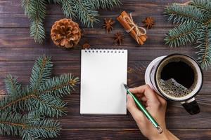 mujer escribe deseos o lista de tareas en cuaderno, taza de café, navidad. ramas de árboles de navidad, piñas, bayas rojas, sobre mesa de mármol, vista superior del espacio de copia foto