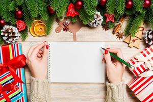 vista superior de la mano femenina escribiendo en un cuaderno sobre fondo de Navidad de madera. abeto y decoraciones festivas. lista de deseos concepto de año nuevo foto