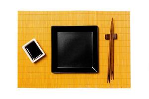 Empty black square plate with chopsticks for sushi and soy sauce on yellow bamboo mat background. Top view with copy space for you design photo