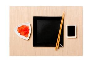 Empty black square plate with chopsticks for sushi, ginger and soy sauce on brown sushi mat background. Top view with copy space for you design photo