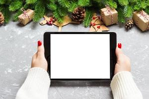 Top view of female hands holding a tablet on cement Christmas background made of fir tree and festive decorations. New year holiday concept. Mockup photo