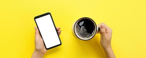 manos femeninas sosteniendo un teléfono móvil negro con pantalla blanca en blanco y una taza de café. imagen de maqueta con espacio de copia para su diseño. banner de vista superior sobre fondo amarillo, plano foto