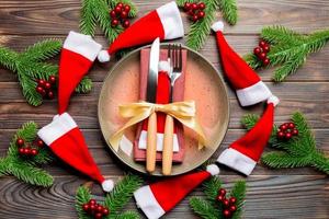 Holiday composition of plate and flatware decorated with Santa hat on wooden background. Top view of Christmas decorations. Festive time concept photo