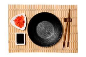 Empty round black plate with chopsticks for sushi and soy sauce, ginger on yellow bamboo mat background. Top view with copy space for you design photo