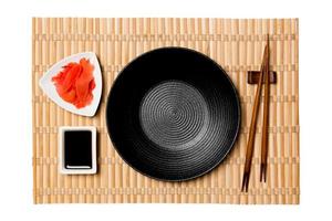 Empty round black plate with chopsticks for sushi and soy sauce, ginger on yellow bamboo mat background. Top view with copy space for you design photo