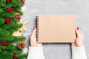 Top view of female hand holding a notebook on cement Christmas background. fir tree and festive decorations. Wish list. New Year concept photo