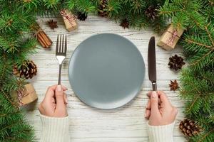 top view girl holds fork and knife in hand and is ready to eat. Empty plate round ceramic on wooden christmas background. holiday dinner dish concept with new year decor photo