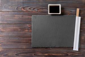 Empty rectangular black slate plate with chopsticks for sushi and soy sauce on wooden background. Top view with copy space photo